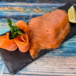 Clare-Island-Räucherlachs in feinen Scheiben auf schwarzer Schieferplatte mit Zitronenspalte angerichtet.