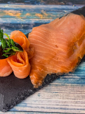 Clare-Island-Räucherlachs in feinen Scheiben auf schwarzer Schieferplatte mit Zitronenspalte angerichtet.