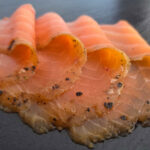 Espresso-Lachs mit dunklem Gewürzrand in dünnen Scheiben auf Schieferplatte angerichtet.