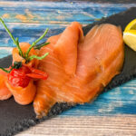 Irischer Wildräucherlachs in feinen Scheiben auf Schieferplatte mit Zitronenspalten angerichtet.