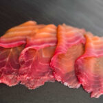 Rote-Bete-Lachs mit rötlichem Rand in dünnen Scheiben auf dunkler Schieferplatte angerichtet.