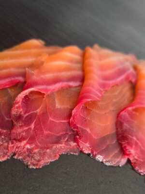 Rote-Bete-Lachs mit rötlichem Rand in dünnen Scheiben auf dunkler Schieferplatte angerichtet.